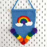 Pennant shape crochet wall hanging in Dolphin Blue with Bright Rainbow and Cloud motif and rainbow pom pom trim, hanging from a wooden dowel and braided hanger. Bright Rainbow Cloud Pennant Wall Hanging by VelvetVolcano