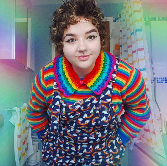 Samantha wearing a rainbow crocheted collar, rainbow striped long sleeve t-shirt, and navy dungarees with rainbow cloud print.
