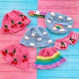 Crochet accessories including 2 pink bucket hats with cherry print, 1 pastel blue bucket hat with cloud print, a pastel rainbow striped bucket hat, a cherry print / striped collar and a light blue & pastel rainbow cloud bum bag