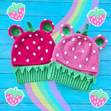 Two VelvetVolcano Strawberry Bear Balaclava's both with Spearmint Green leaves and neck ribbing. The one on the left is hot pink and the one on the right is bubblegum pink. They both feature white polka dot seeds.