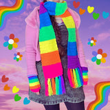 A woman's torso and upper legs are in frame, she is wearing a grey/black acid wash denim pinafore dress, a rainbow striped top with a heart cut out on the chest, a pink ribbed cardigan with a faux fur collar, black tights and a matching neon rainbow striped crochet scarf with rainbow tassels and neon rainbow striped crochet fingerless gloves that are by VelvetVolcano