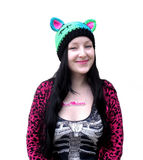 Tamsyn, a white woman with long black hair is wearing a VelvetVolcano FrankenKitty Beanie which is a crochet cat ear hat, half turquoise and half neon green with black stitch details inspired by Frankenstein's Monster