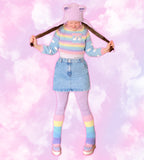 Tamsyn, is standing with her feet pointed inwards, holding the ends of her long hair (styled in bunches) in her fists & pulling it outwards in a cute pose. She’s wearing a pastel outfit with 3 crocheted pieces - a lilac fuzzy earflap hat with bear ears, a cropped pastel rainbow striped sweater with cloud print sleeves & a pair of pastel rainbow striped flared leg warmers. The other pieces in the outfit are lilac leggings, a high waist light denim mini skirt and a pair of pastel platform boots