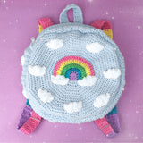 Light blue crochet circular backpack with pastel rainbow striped straps and a pastel rainbow and cloud design on the front by VelvetVolcano