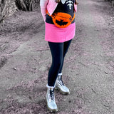 A photo of Tamsyn wearing a white and pink ombre biker jacket, a black sweater with white skeleton rib print, a pink corduroy mini skirt, black leggings, silver Dr Martens boots and an orange Creature Feature Bum Bag by VelvetVolcano. The background is a spooky looking wooded area. Tamsyn's head is out of frame but her brown hair can be seen in plaits.