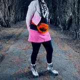 A photo of Tamsyn wearing a white and pink ombre biker jacket, a black sweater with white skeleton rib print, a pink corduroy mini skirt, black leggings, silver Dr Martens boots and an orange Creature Feature Bum Bag by VelvetVolcano. The background is a spooky looking wooded area. Tamsyn's head is out of frame but her brown hair can be seen in plaits.