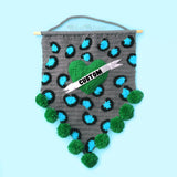 Grey crochet pennant wall hanging with turquoise and black leopard print and a green love heart with a white felt banner over the top that says "Custom" in black text. The triangular bottom of the wall hanging is trimmed with green pom poms and it hangs from a wooden dowel.