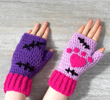 VelvetVolcano NecroKitty / FrankenKitty crochet fingerless gloves. One glove is violet, the other is lilac. Both feature hot pink heart shaped paw prints, black Frankenstein's Monster inspired stitches and hot pink cuffs.