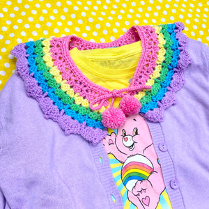 Kawaii Pastel Rainbow Striped Detachable Crocheted Collar - Bubblegum Pink, Pastel Yellow, Light Green, Turquoise and Lilac Striped Peter Pan Collar with Pom Pom Ties by VelvetVolcano shown on top of a lilac cardigan and a yellow t-shirt with and image of Cheer Bear the Care Bear on it. The background is yellow with white polka dots.