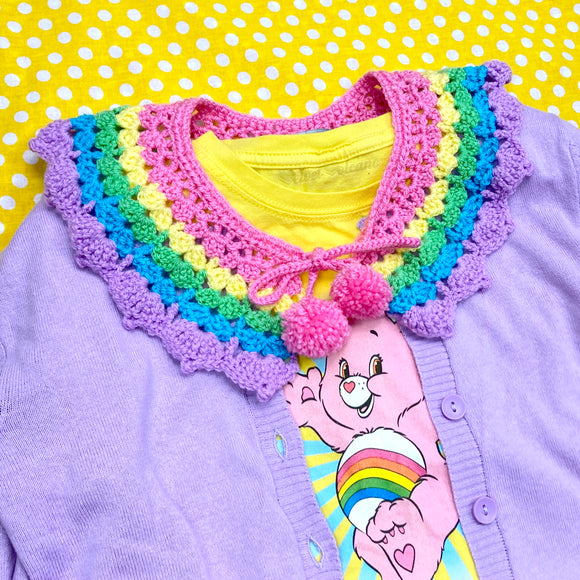 Kawaii Pastel Rainbow Striped Detachable Crocheted Collar - Bubblegum Pink, Pastel Yellow, Light Green, Turquoise and Lilac Striped Peter Pan Collar with Pom Pom Ties by VelvetVolcano shown on top of a lilac cardigan and a yellow t-shirt with and image of Cheer Bear the Care Bear on it. The background is yellow with white polka dots.