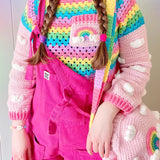 Tamsyn (a white woman in her early 30's with long plaited brown hair) is wearing a VelvetVolcano pastel rainbow outfit with cropped crochet sweater, circular shoulder bag and beret and hot pink Lucy & Yak dungarees