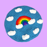 A dolphin blue crocheted beret with a repeating pattern of white clouds and a bright rainbow in the centre with white clouds at both ends by VelvetVolcano