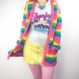 A woman wearing a VelvetVolcano Pastel Rainbow Striped Crochet Cardigan, a sparkly pastel rainbow heart necklace, a graphic tee, a pastel yellow mini-skirt and pastel pink leggings.