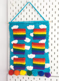 Turquoise crochet wall hanging organiser with white cloud pattern and 6 bright rainbow striped pockets. The wall hanging hangs from a wooden dowel and braided hanger and the bottom is trimmed with 7 pom poms, each in a different rainbow colour. Clouds & Rainbows Hanging Organiser by VelvetVolcano